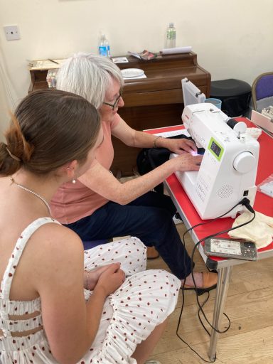 An older woman demonstrates sewing techniques to a younger woman at a sewing machine.