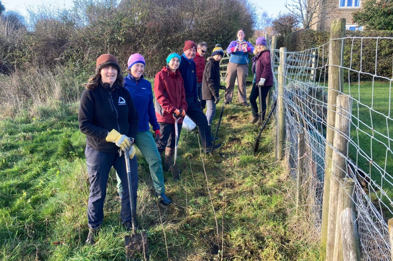 Five people in red and black jackets gardening, digging in a green area.