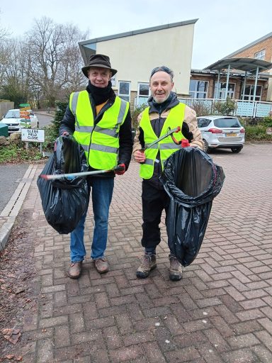 Two men in reflective vests holding black rubbish bags, standing in a car park.