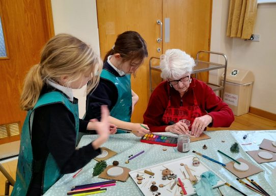 Two children and an elderly woman engaged in a creative craft activity at a table.